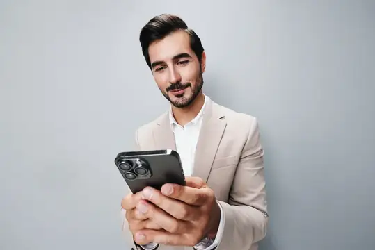 Улыбающийся бородатый мужчина в бежевом пиджаке смотрит в смартфон