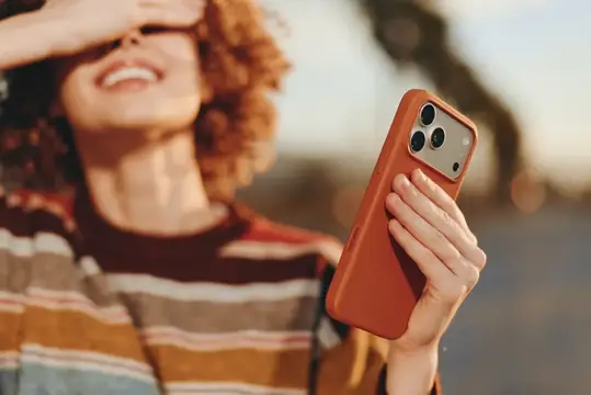 Рыжеволосая женщина смеётся, держа смартфон в терракотовом чехле