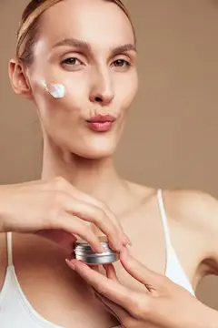 Женщина, уход за кожей, красота, крупный план, крем для лица на щеке, руки держат баночку, нежный уход для гладкой кожи, мягкий свет, расслабленная оздоровительная рутина, бежевая студия…