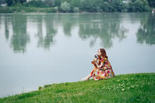 Женщина в платье с фотоаппаратом в руках стоит на берегу озера