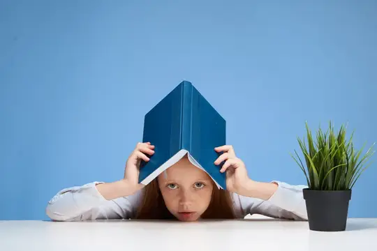 Рыжеволосая девочка спряталась под синей книгой за столом