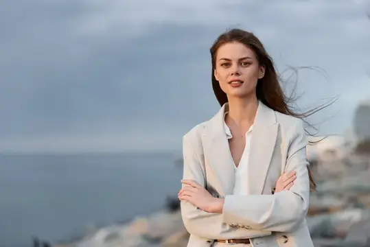 Молодая женщина в светлом пиджаке стоит у моря на ветру