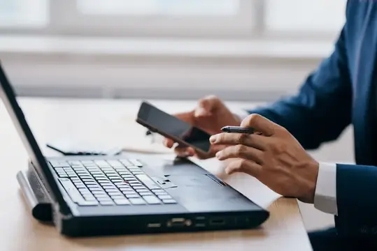 Мужчина в костюме работает за ноутбуком и держит смартфон
