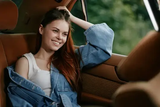 Улыбающаяся рыжеволосая девушка в джинсовой куртке сидит в салоне автомобиля