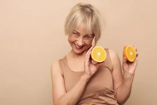 Счастливая зрелая женщина с blond-волосами в бежевом топе держит половинки апельсинов близко к лицу на однотонном фоне