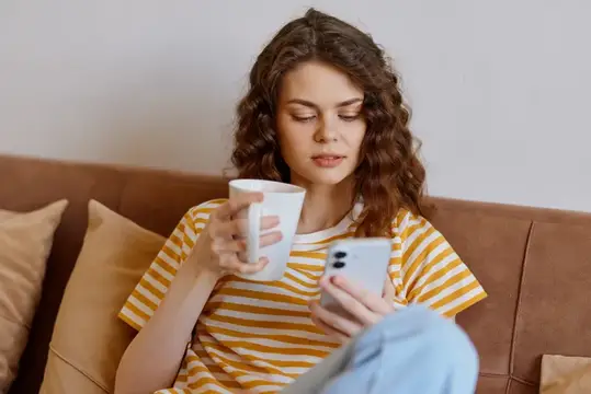 Кудрявая девушка с кружкой смотрит в смартфон на диване