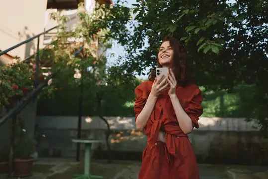 Улыбающаяся темноволосая девушка в красном платье смотрит в телефон в саду