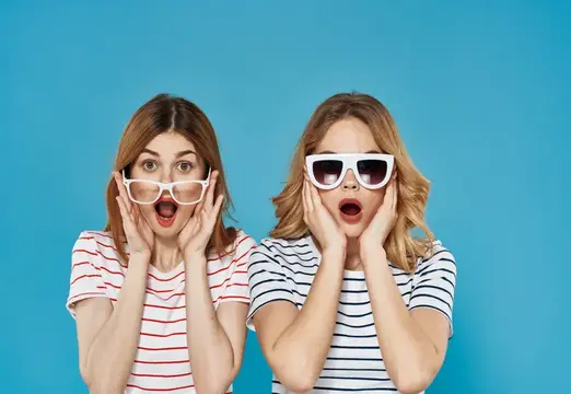 Две удивлённые девушки в очках хватаются за щёки на синем фоне