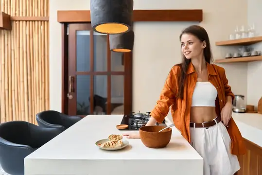 Молодая женщина в оранжевой куртке стоит на современной кухне и улыбается, готовя здоровую пищу