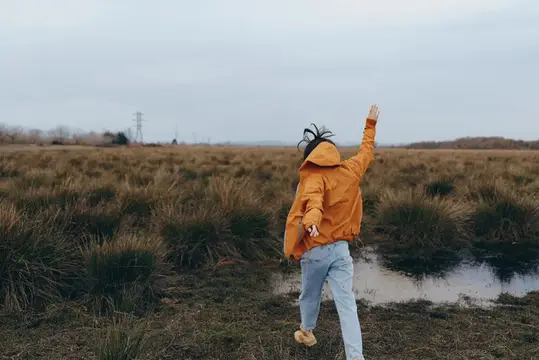 Темнокожая девушка в жёлтом плаще идёт по осеннему полю с кочками