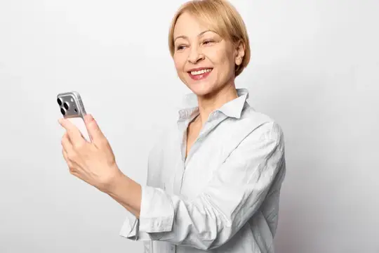 Улыбающаяся женщина со светлыми волосами смотрит в смартфон на светлом фоне