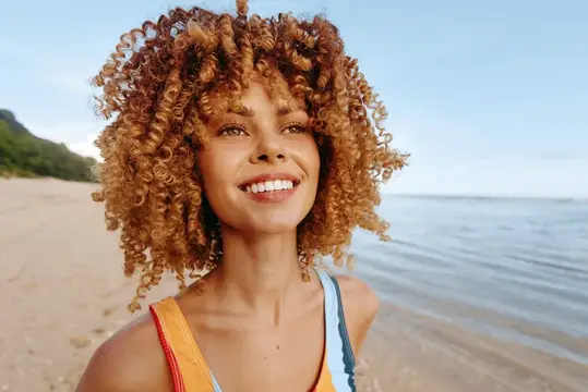 Улыбающаяся темнокожая девушка с рыжими кудрями на пляже у моря