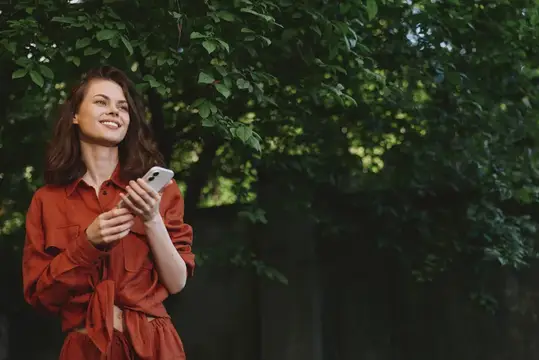 Улыбающаяся темноволосая женщина с телефоном в красной рубашке у деревьев