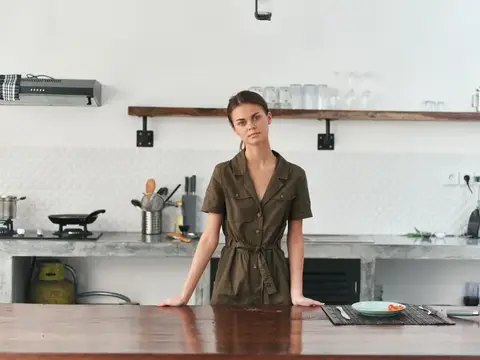 Молодая женщина в оливковом платье стоит у кухонного стола