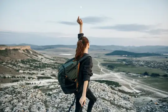 Торжествующая женщина на горной вершине с рюкзаком тянется руками к небу, празднуя свою победу