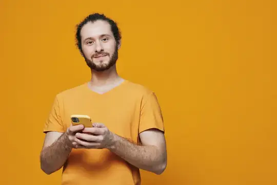 Молодой бородатый мужчина в оранжевой футболке с телефоном на оранжевом фоне