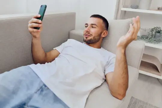Молодой мужчина лежит на диване со смартфоном и жестикулирует