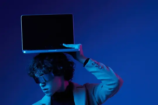 Молодой человек в VR-очках держит ноутбук на голове в синем свете