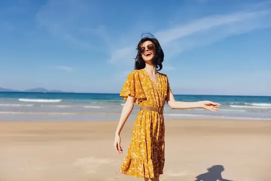 Улыбающаяся женщина в жёлтом платье гуляет по пляжу у моря