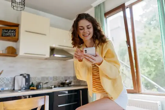 Яркая сцена улыбающейся женщины дома, на кухне, использующей смартфон, повседневная одежда, легкий ветерок из окна, спокойное настроение, простой лайфстайл-момент