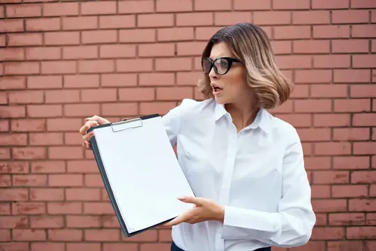 Удивлённая женщина в очках с планшетом на фоне кирпичной стены