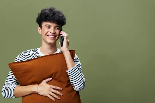 Улыбающийся кудрявый парень с подушкой говорит по телефону