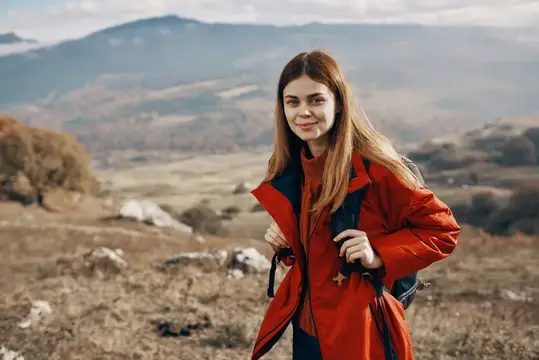 Улыбающаяся девушка в красной куртке с рюкзаком на фоне гор
