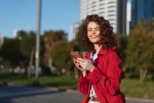 Улыбающаяся кудрявая девушка в красном плаще с телефоном на городской улице