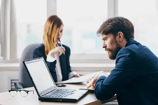 Бородатый мужчина в офисе работает за ноутбуком на фоне коллеги