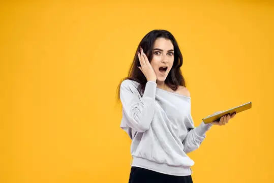Удивлённая брюнетка с книгой прикрывает рот рукой на жёлтом фоне