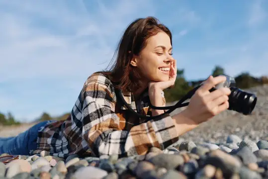 Женщина-фотограф отдыхает на камнях у пляжа, с радостным выражением лица, снимает сцены фотоаппаратом, одета в клетчатую рубашку