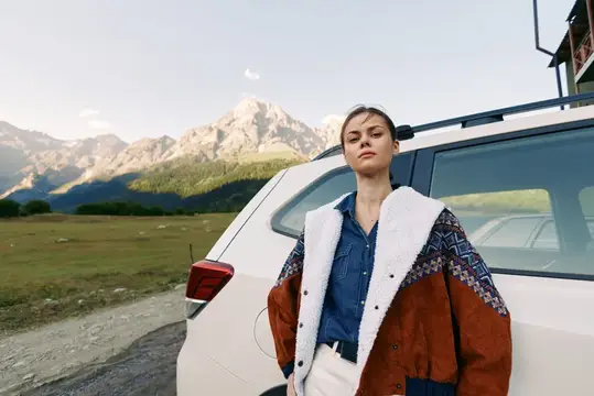Молодая женщина в куртке с белым воротником стоит у внедорожника на фоне гор