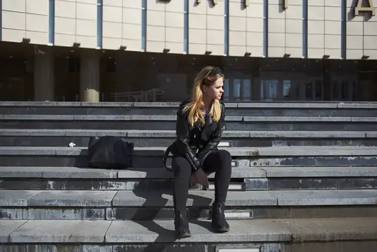 Блондинка в кожанке сидит на ступенях у здания