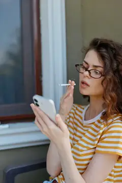 Непринужденная женщина на открытом воздухе пользуется телефоном, в руке сигарета, в очках, в полосатой футболке