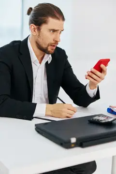 Мужчина в костюме удивлённо смотрит на красный смартфон за рабочим столом