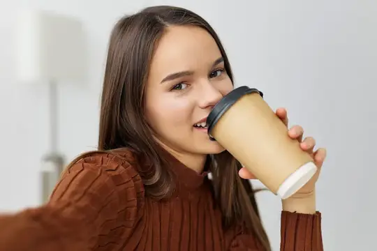Улыбающаяся девушка с кофе в бумажном стакане смотрит в камеру