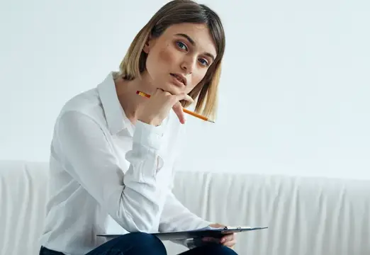 Женщина сидит на диване в светлой комнате, на ней рубашка, а на коленях лежат бумаги