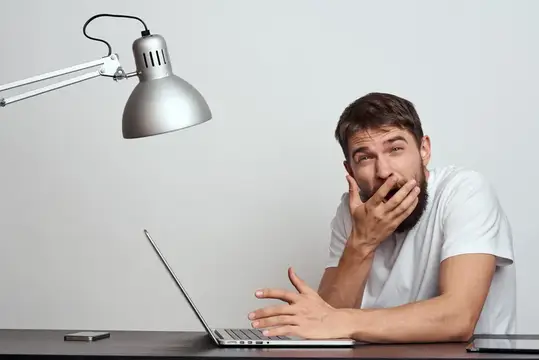 Бородатый мужчина в белой футболке удивлённо прикрывает рот рукой за ноутбуком