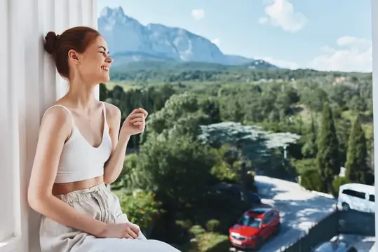 Рыжеволосая девушка у окна любуется горами и лесом