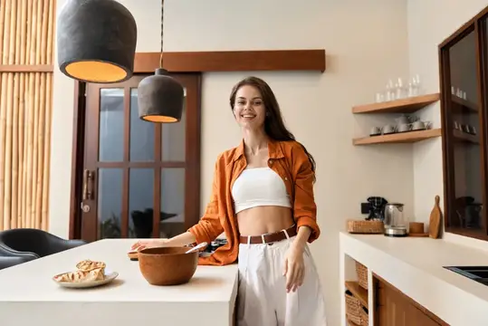 Улыбающаяся девушка в оранжевой рубашке готовит на светлой кухне