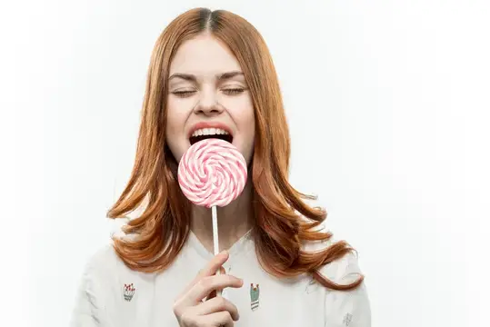 Рыжеволосая девушка с удовольствием кусает большую круглую конфету на палочке
