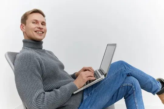 Улыбающийся мужчина в свитере работает за ноутбуком в кресле
