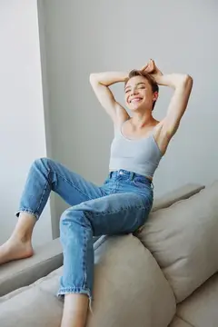 Молодая женщина в джинсах и майке потягивается на диване с улыбкой