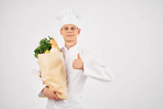 Молодой повар в колпаке с пакетом продуктов показывает большой палец вверх