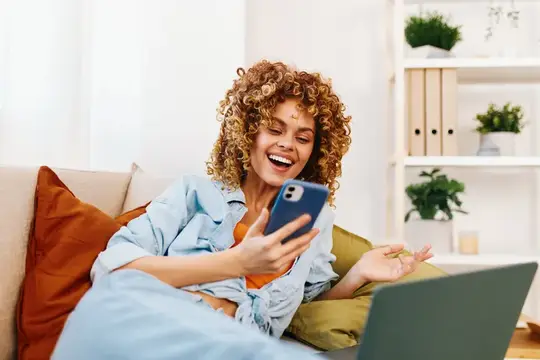 Кудрявая девушка смеётся, глядя в смартфон, лёжа на диване