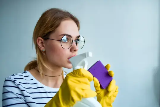 Молодая женщина в очках и жёлтых перчатках с чистящим средством и губкой