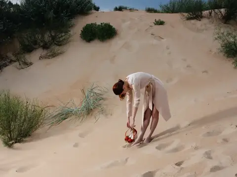 Босая женщина в белом платье наклоняется с корзиной на песчаной дюне
