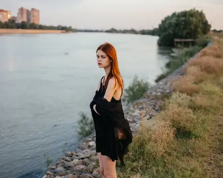 Рыжеволосая женщина в черном платье стоит у реки на природе