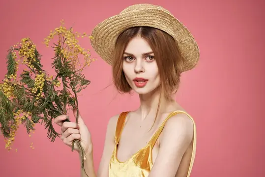 Молодая женщина в соломенной шляпе с веткой мимозы на розовом фоне