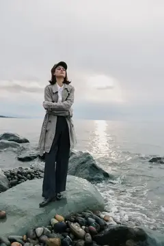 Молодая женщина в плаще и кепке стоит на камнях у моря